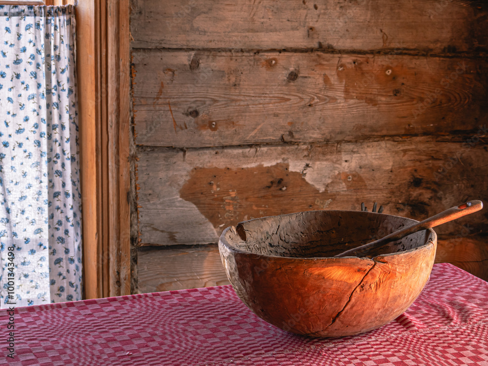 In a rustic house in the kitchen, there is an old wooden bowl with a spoon on the table. The interior of the kitchen of a rural house built in the 19th century. A red tablecloth on an antique table. In a rustic house in the kitchen, there is an old wooden bowl with a spoon on the table. The interior of the kitchen of a rural house built in the 19th century. A red tablecloth on an antique table.