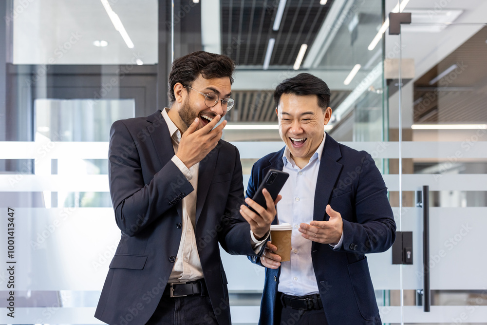 Two colleagues sharing a light moment in office while watching a funny video on smartphone. Both men dressed in business suits, one holding a coffee cup, expressing laughter and surprise. Two colleagues sharing a light moment in office while watching a funny video on smartphone. Both men dressed in business suits, one holding a coffee cup, expressing laughter and surprise.