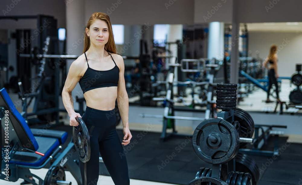 Young woman lifting weights in a gym setting. A young woman stands confidently in a gym, holding a weight plate, showcasing strength during her workout session. Young woman lifting weights in a gym setting. A young woman stands confidently in a gym, holding a weight plate, showcasing strength during her workout session.