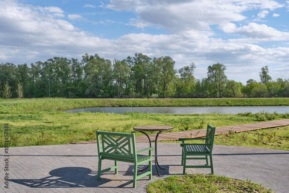 Serene outdoor scene in park. Two green wooden chairs, round table. Calm pond, surrounded by trees. Serene outdoor scene in park. Two green wooden chairs, round table. Calm pond, surrounded by trees.