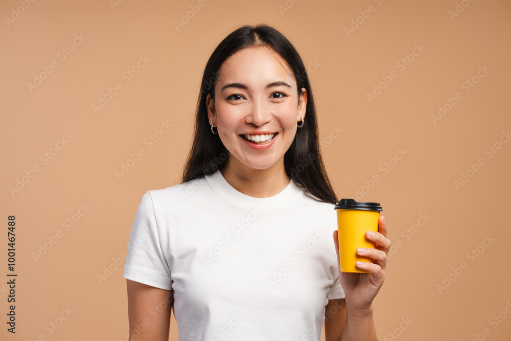 Smiling asian woman holding takeaway coffee cup posing in studio on beige background Smiling asian woman holding takeaway coffee cup posing in studio on beige background