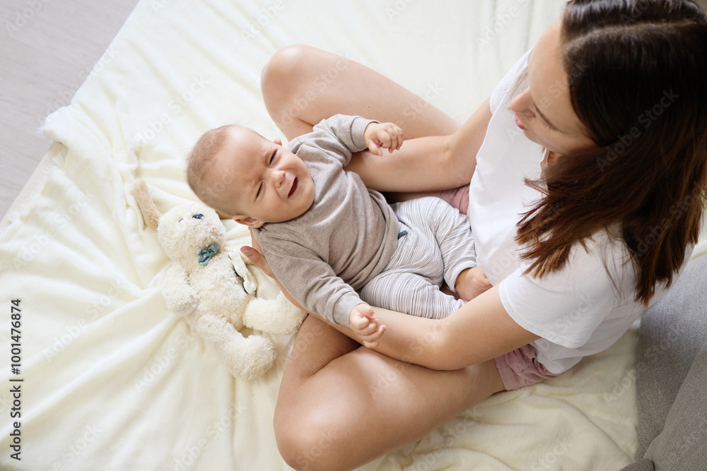 Loving parent sitting on bed at home holding her little infant crying boy close comforting him with gentle touches and soothing words to calm down during tender family bonding moment Loving parent sitting on bed at home holding her little infant crying boy close comforting him with gentle touches and soothing words to calm down during tender family bonding moment