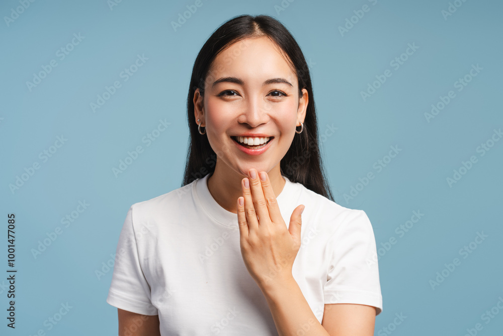 Smiling asian woman is laughing and covering her mouth with hand closeup isolated on blue background Smiling asian woman is laughing and covering her mouth with hand closeup isolated on blue background