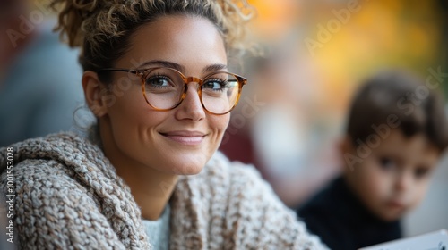 A woman with curly hair and glasses smiles warmly while enjoying an outdoor setting, with a blurred child in the background conveying happiness.