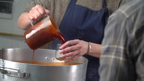A worker in a jam factory pours freshly prepared Rosa Mosqueta (rosehip) jam into small glass jars. The production takes place in an artisanal setting, emphasizing the handcrafted quality of the proce
