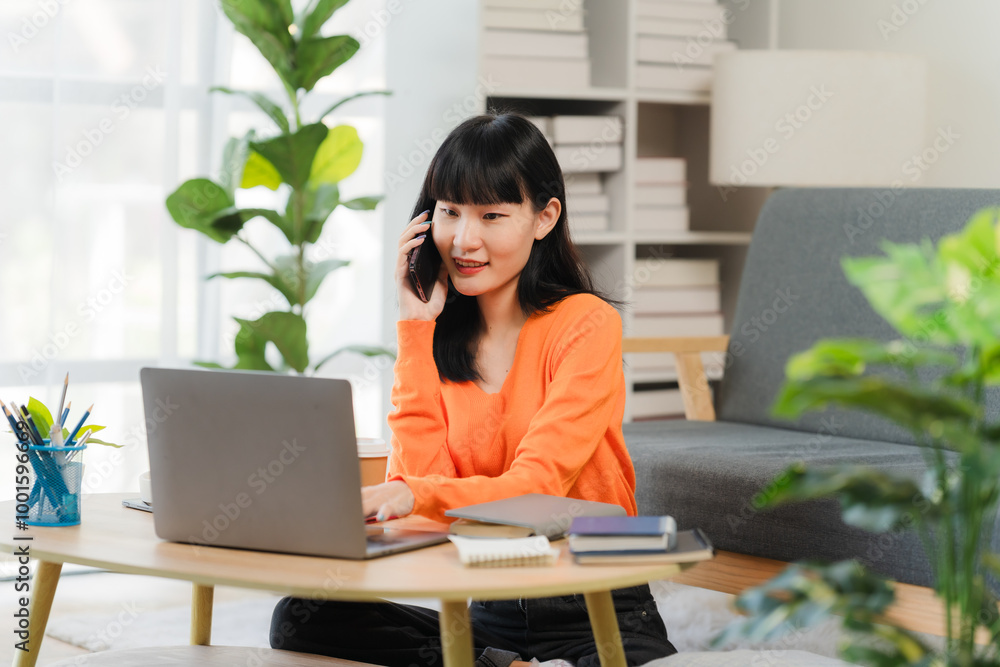 Focused and Connected: Young Asian woman working from home, effortlessly balancing a phone call and laptop work. A vibrant image of modern remote work life. Focused and Connected: Young Asian woman working from home, effortlessly balancing a phone call and laptop work. A vibrant image of modern remote work life.