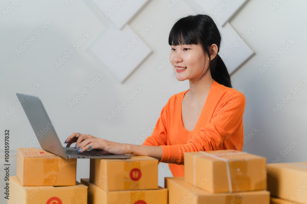 Online Business Owner: Happy Asian woman working on laptop, surrounded by packages.  E-commerce success story. 