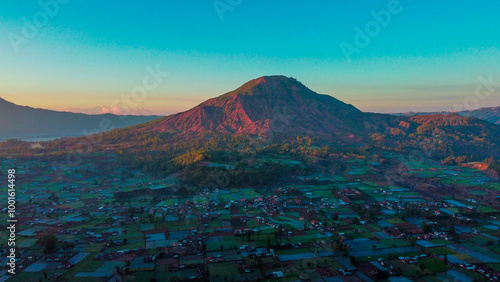 A stunning sunset over Mount Batur illuminates lush rice fields and cozy villages, bathed in soft morning mist, creating a serene.