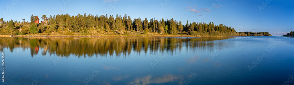 Wide-angle panorama of a tranquil sea with reflections of a dense forest and distant red lighthouse under a clear blue sky, creating a peaceful atmosphere. Wide-angle panorama of a tranquil sea with reflections of a dense forest and distant red lighthouse under a clear blue sky, creating a peaceful atmosphere.