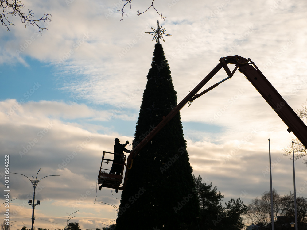 A silhouette of a man decorating a high artificial holiday tree from crane truck in Peremohy Park in Kyiv, Ukraine. Festive December preparations for Christmas and New Year.