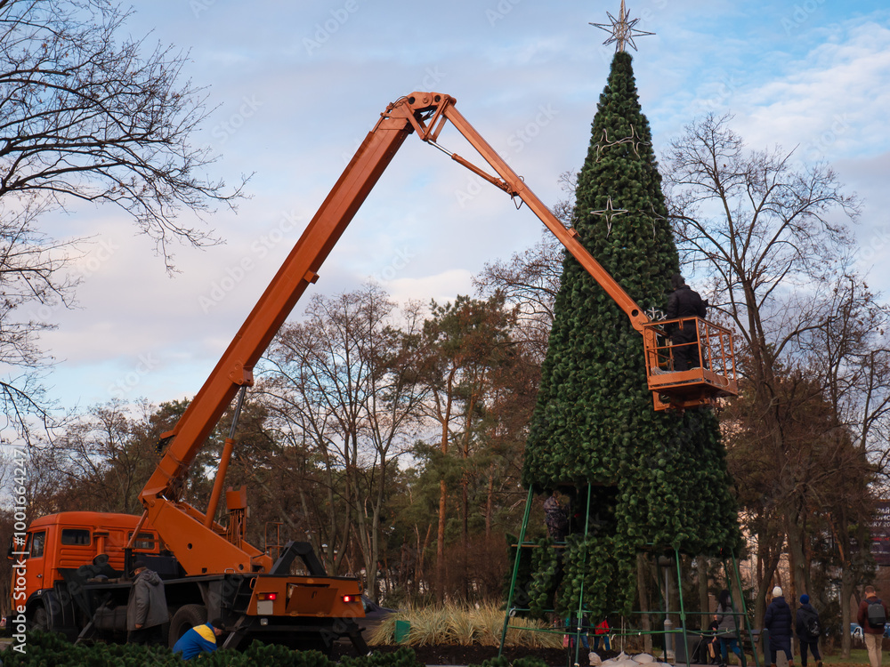 People on an orange crane truck decorating a high artificial holiday tree in Peremohy Park in Kyiv, Ukraine. Festive December preparations for Christmas and New Year.