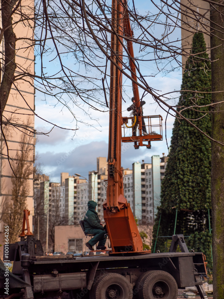 Blurry view of people decorating a high artificial holiday tree with the help of an orange crane truck in Peremohy Park in Kyiv, Ukraine. Festive December preparations for Christmas and New Year.