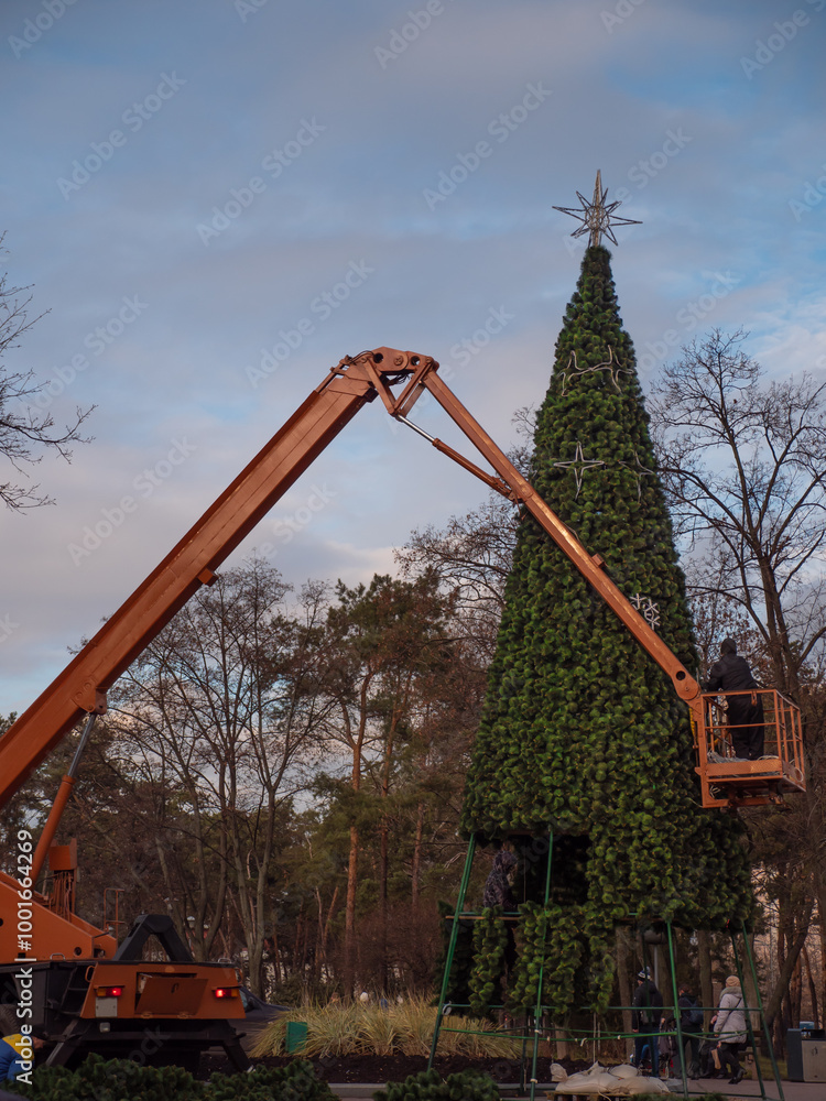 People on an orange crane truck decorating a high artificial holiday tree in Peremohy Park in Kyiv, Ukraine. Festive December preparations for Christmas and New Year.