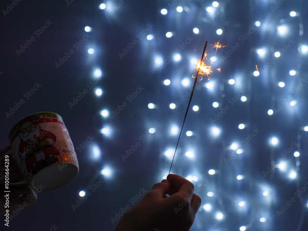Female hand holding sparkling yellow Christmas bengali light in one hand and a cup in another. String fairy lights glowing on blue background. Christmas and New Year celebration concept.
