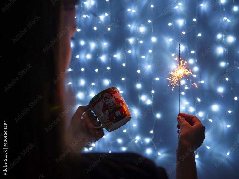 Girl holding sparkling yellow Christmas bengali light in one hand and a cup in another. String fairy lights glowing on blue background. Christmas and New Year concept.