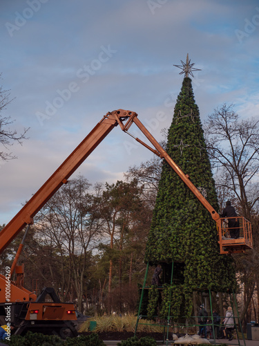 Obraz na płótnie People on an orange crane truck decorating a high artificial holiday tree in Peremohy Park in Kyiv, Ukraine. Festive December preparations for Christmas and New Year. w salonie