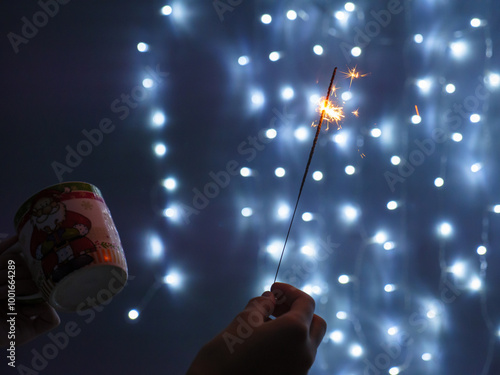 Obraz na płótnie Female hand holding sparkling yellow Christmas bengali light in one hand and a cup in another. String fairy lights glowing on blue background. Christmas and New Year celebration concept. w salonie