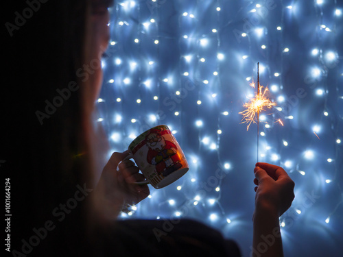Obraz na płótnie Girl holding sparkling yellow Christmas bengali light in one hand and a cup in another. String fairy lights glowing on blue background. Christmas and New Year concept. w salonie