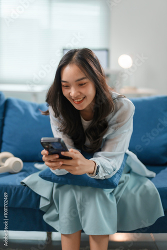 Young woman is sitting on a blue couch at home, smiling while using a smartphone