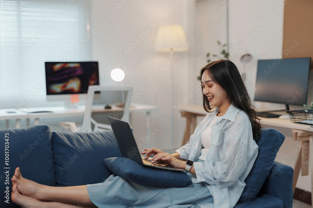 Smiling asian woman working remotely, sitting comfortably on the sofa in her living room, using laptop