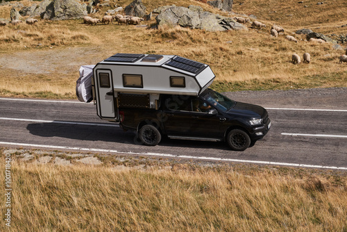 Black pickup truck with a pop-up camper driving on a winding mountain road. The scenic landscape is a mix of green trees, rocky hills, and dry grass. Carpathian Mountains