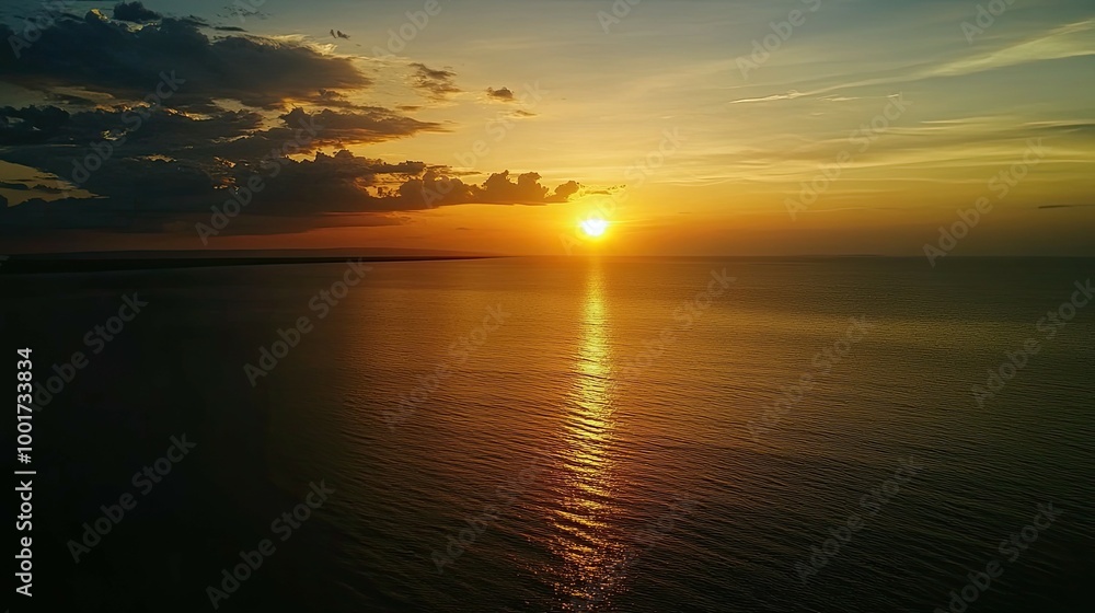 Naklejka premium A stunning sunset over the ocean with a bright sun and golden light reflected on the water. The sky is a mix of orange, yellow, and blue with dramatic clouds.
