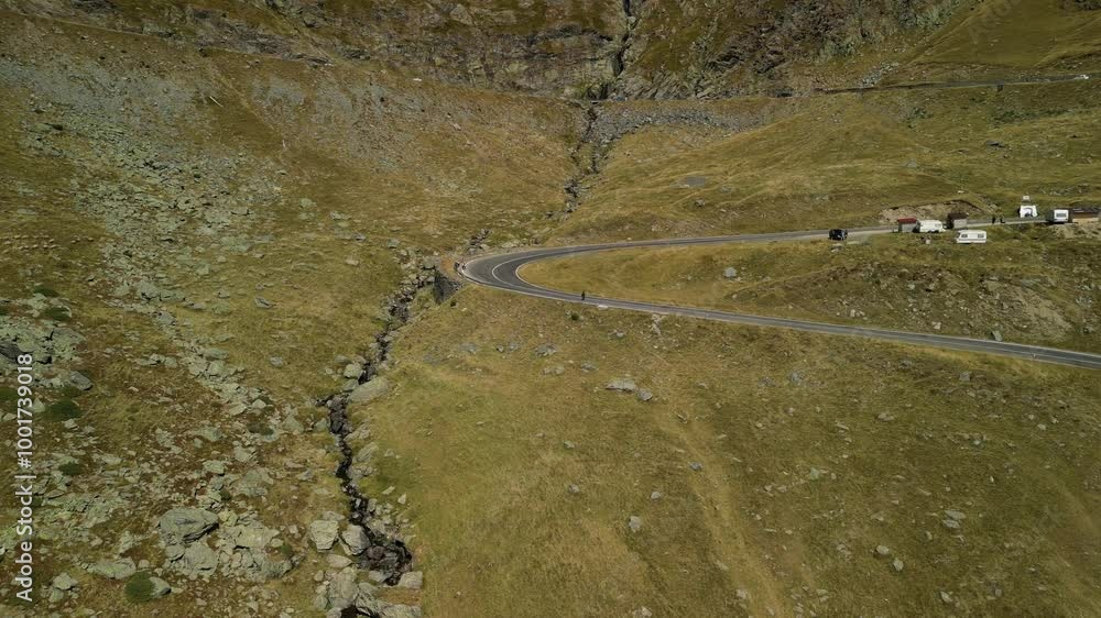 Aerial view of a winding road in the mountains. The road curves through a grassy valley, with a small stream running alongside it. Transfăgărășan road, Romania Aerial view of a winding road in the mountains. The road curves through a grassy valley, with a small stream running alongside it. Transfăgărășan road, Romania
