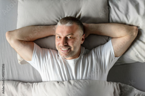 Relaxed European middle aged man laying in comfy bed with hands behind head and smiling at camera, enjoying new day, top view shot