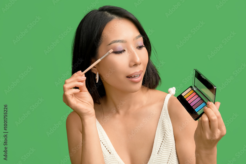 Young Asian woman with palette of eyeshadows and makeup brush on green background Young Asian woman with palette of eyeshadows and makeup brush on green background