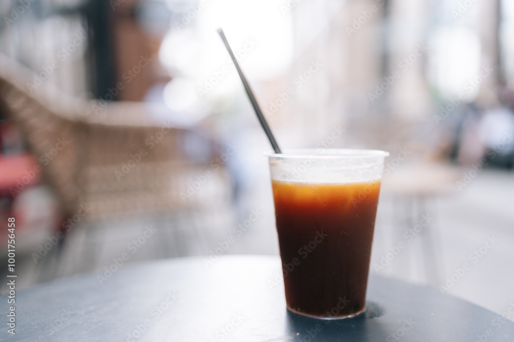 Close-up front view of plastic cup of iced coffee with black straw sitting on table at outdoor cafe with blurry background of buildings, no people. Concept of drink, lifestyle, city life Close-up front view of plastic cup of iced coffee with black straw sitting on table at outdoor cafe with blurry background of buildings, no people. Concept of drink, lifestyle, city life