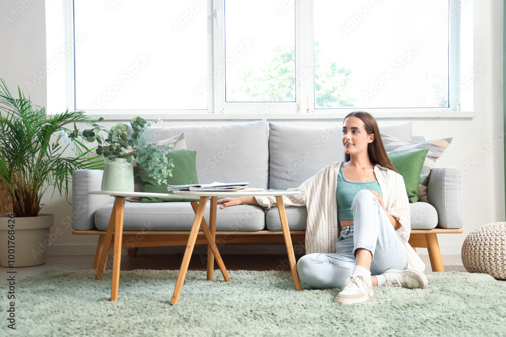 Young woman sitting on green carpet near grey sofa in living room Young woman sitting on green carpet near grey sofa in living room