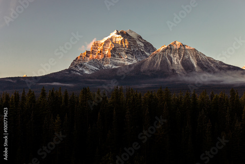 Fotografie A breathtaking sunset casts a warm glow on the majestic snowy peaks, highlighting the forest covers below