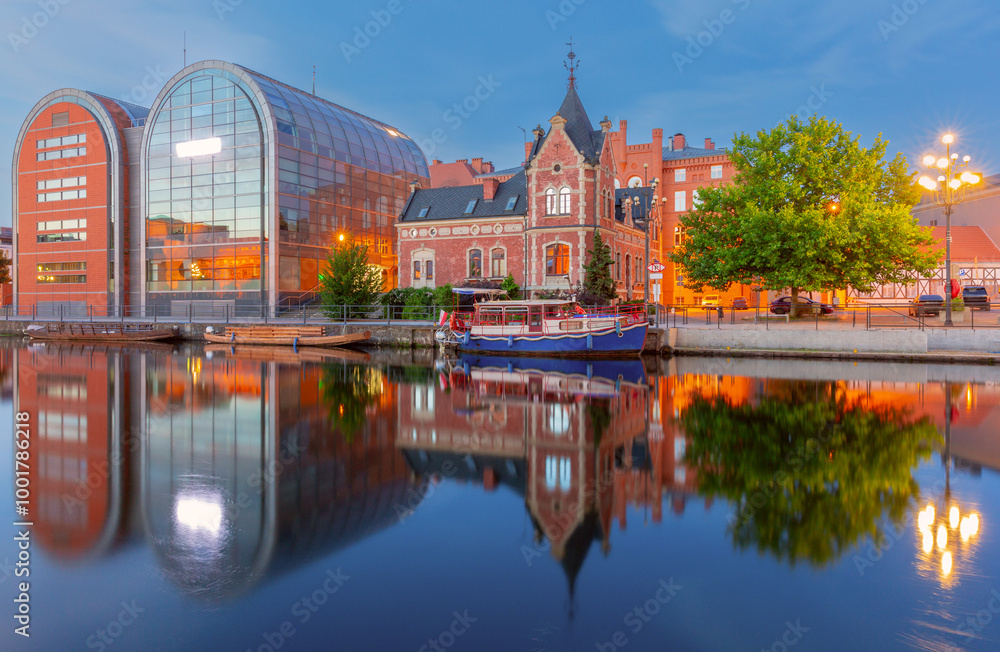 Fototapeta premium View of the Bydgoszcz waterfront featuring a modern glass building and a historic red brick structure, with reflections in the Brda River in Poland