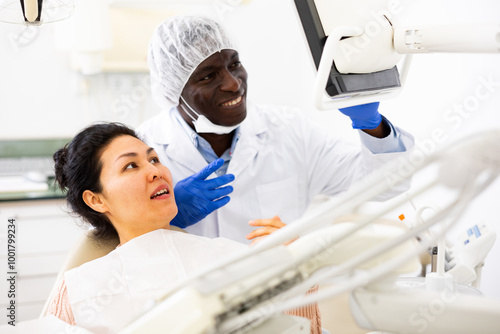 Positive african american dentist talking to asian woman during dental checkup, discussing teeth x-ray and treatment procedures photo