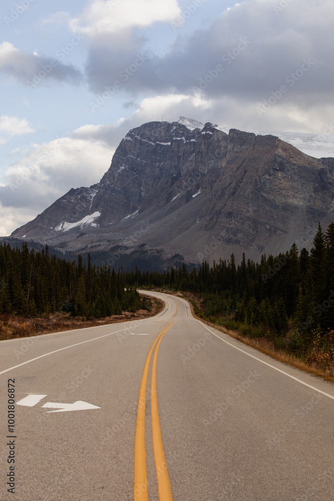 view of a curve road in a pine tree forest with a tall rocky mountain in the distance with clouds in the sky in fall view of a curve road in a pine tree forest with a tall rocky mountain in the distance with clouds in the sky in fall