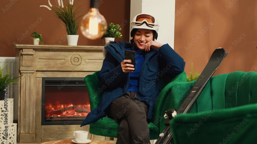 Asian woman laughing at smartphone and talking to friends about extreme winter sport with skis, preparing to go sliding on mountain alpine slopes. Young adult checking online websites. Handheld shot. Asian woman laughing at smartphone and talking to friends about extreme winter sport with skis, preparing to go sliding on mountain alpine slopes. Young adult checking online websites. Handheld shot.