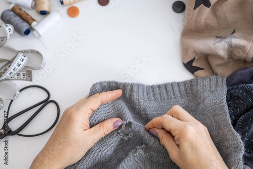 A woman uses her hands to sew up a hole in a gray knitted garment with a needle and thread. 