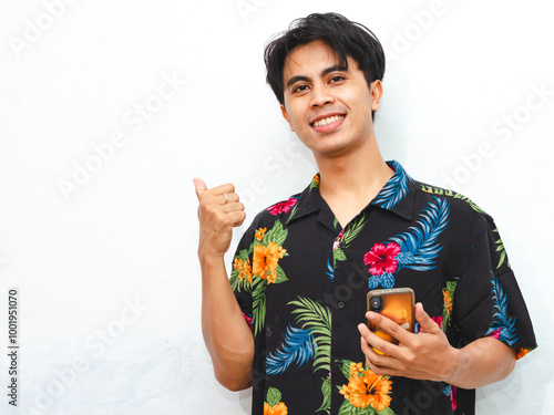 Young Asian man in casual summer clothes holding a smartphone, pointing with his thumb, presenting an ad with a confident expression. photo