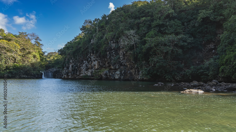 Fototapeta premium Ocean Bay. There is green tropical vegetation on the steep rocky shores. A waterfall is visible in the distance. Streams flow into the sea. Blue sky, clouds. Mauritius.Grand River South East waterfall