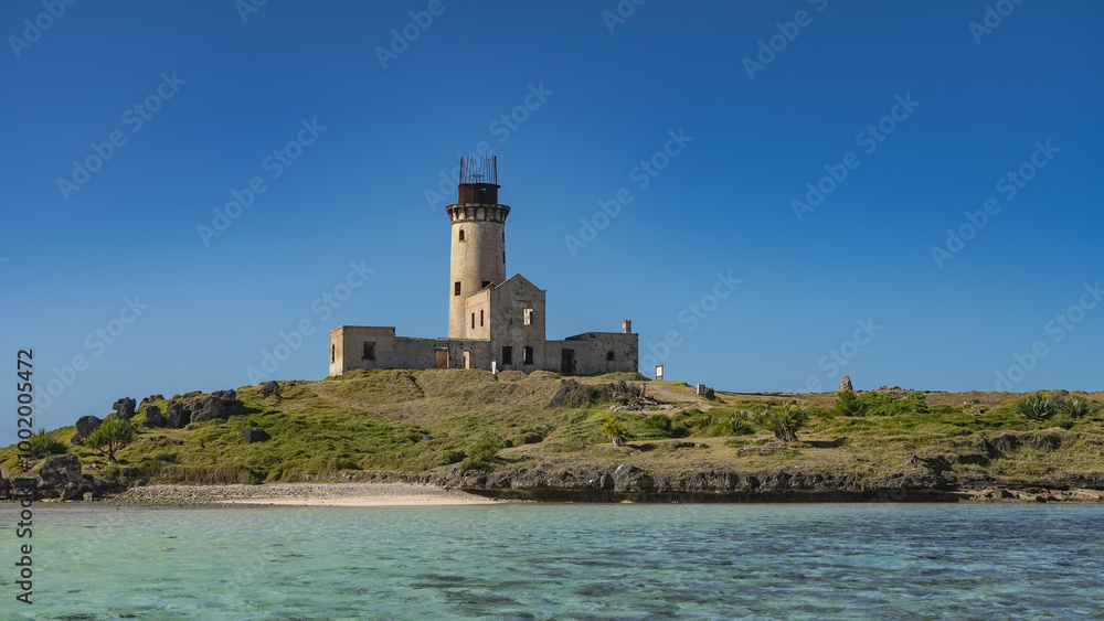 An old lighthouse on a tropical island. A dilapidated building, a tall cylindrical tower against a clear blue sky. Green vegetation all around. In the foreground is an aquamarine ocean. Mauritius. An old lighthouse on a tropical island. A dilapidated building, a tall cylindrical tower against a clear blue sky. Green vegetation all around. In the foreground is an aquamarine ocean. Mauritius.