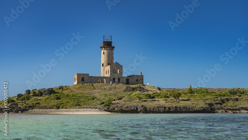 An old lighthouse on a tropical island. A dilapidated building, a tall cylindrical tower against a clear blue sky. Green vegetation all around. In the foreground is an aquamarine ocean. Mauritius.