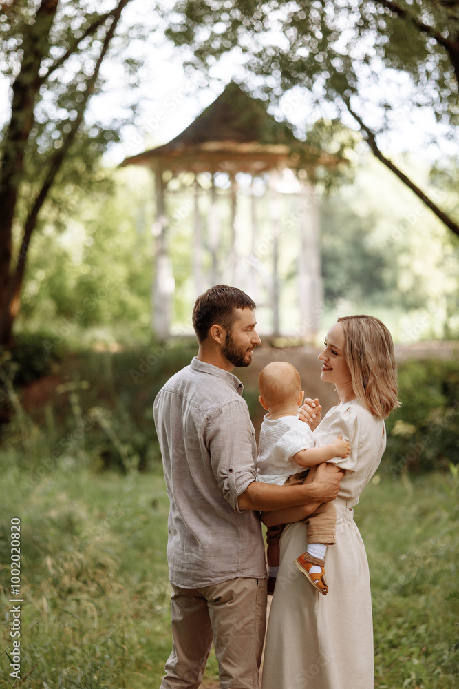 A family of three, a man and a woman with a child in their arms, stand in a park in the summer. The concept of a happy family with a small child, care and love. A family of three, a man and a woman with a child in their arms, stand in a park in the summer. The concept of a happy family with a small child, care and love.