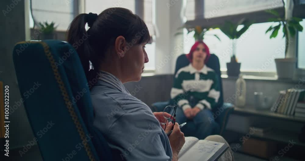 Back view of female therapist with notebook sitting in chair, providing therapy for teenager. Teenage girl with pink hair shares feelings, discusses problems with psychologist on counseling session. Back view of female therapist with notebook sitting in chair, providing therapy for teenager. Teenage girl with pink hair shares feelings, discusses problems with psychologist on counseling session.