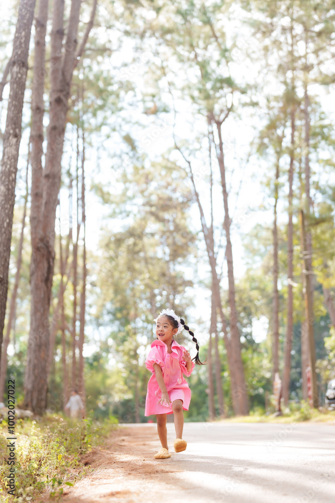 Asian girl running energetically down a park path wearing a pink dress and white headband. Child enjoying freedom and playfulness in nature, surrounded by trees and a sunny environment. Asian girl running energetically down a park path wearing a pink dress and white headband. Child enjoying freedom and playfulness in nature, surrounded by trees and a sunny environment.