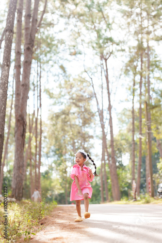 Asian girl running energetically down a park path wearing a pink dress and white headband. Child enjoying freedom and playfulness in nature, surrounded by trees and a sunny environment.