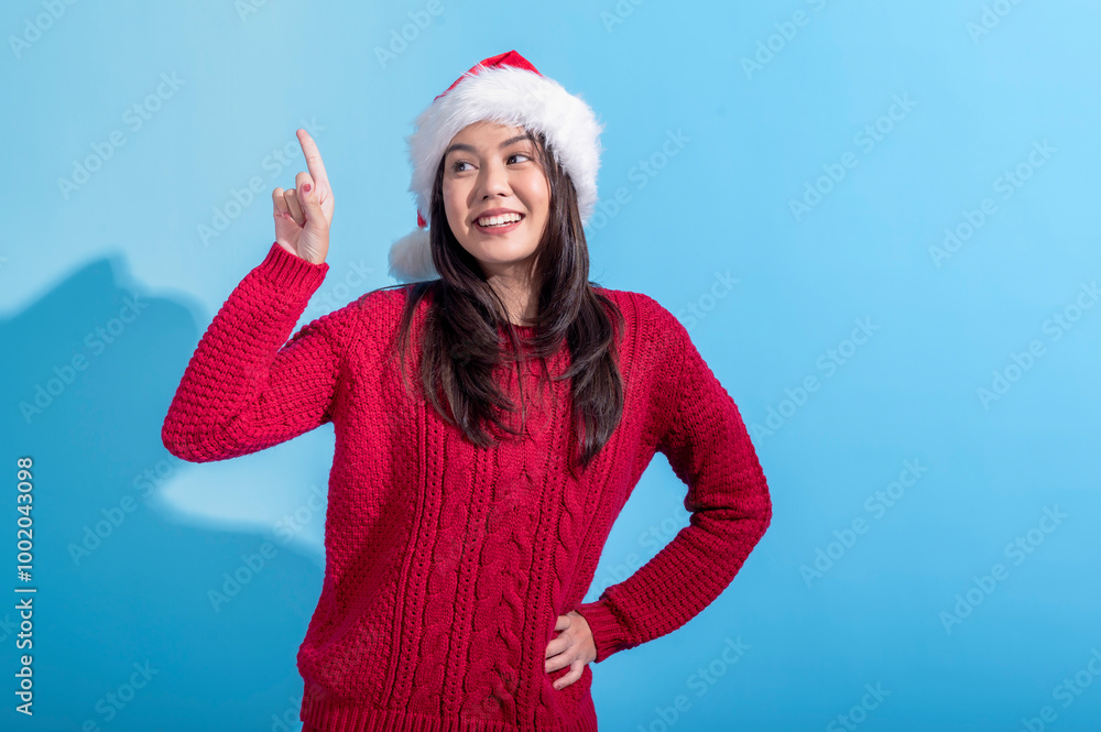 An Asian woman in a red knit sweater and Santa hat poses with an enthusiastic expression. She points upward with one finger while smiling. The background is a solid light blue color, An Asian woman in a red knit sweater and Santa hat poses with an enthusiastic expression. She points upward with one finger while smiling. The background is a solid light blue color,