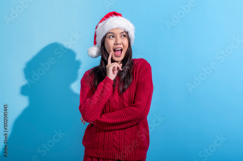 An Asian woman in a red knit sweater and Santa hat strikes a playful, surprised pose with her finger on her chin. She stands against a light blue background, exuding joyful Christmas vibes.