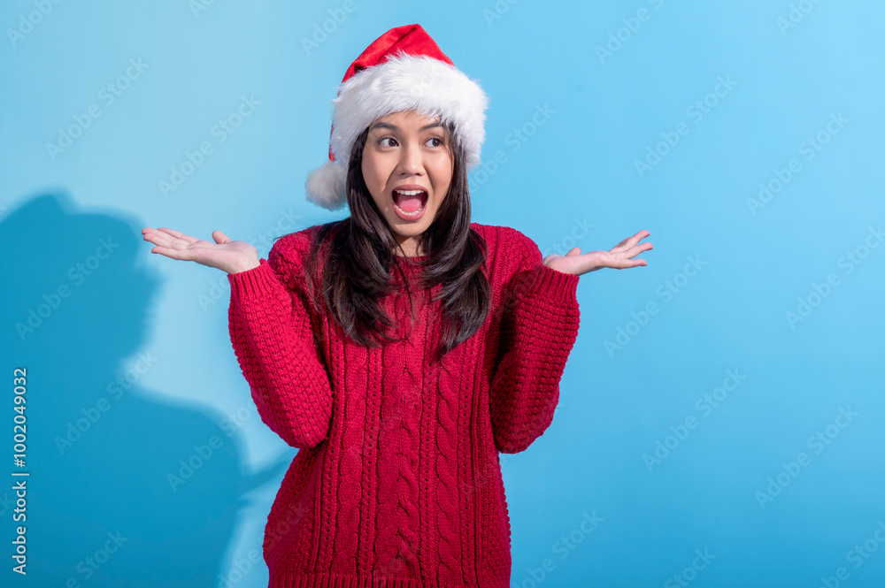 Obraz premium An Asian woman wearing a red knit sweater and a Santa hat stands with her hands raised in a surprised. Her mouth is open in an expressive. The background is light blue.
