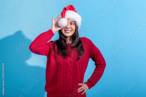 An Asian woman in a red sweater and Santa hat playfully covers one eye with the fluffy white pompom of her hat, smiling cheerfully. She is posing against a light blue background