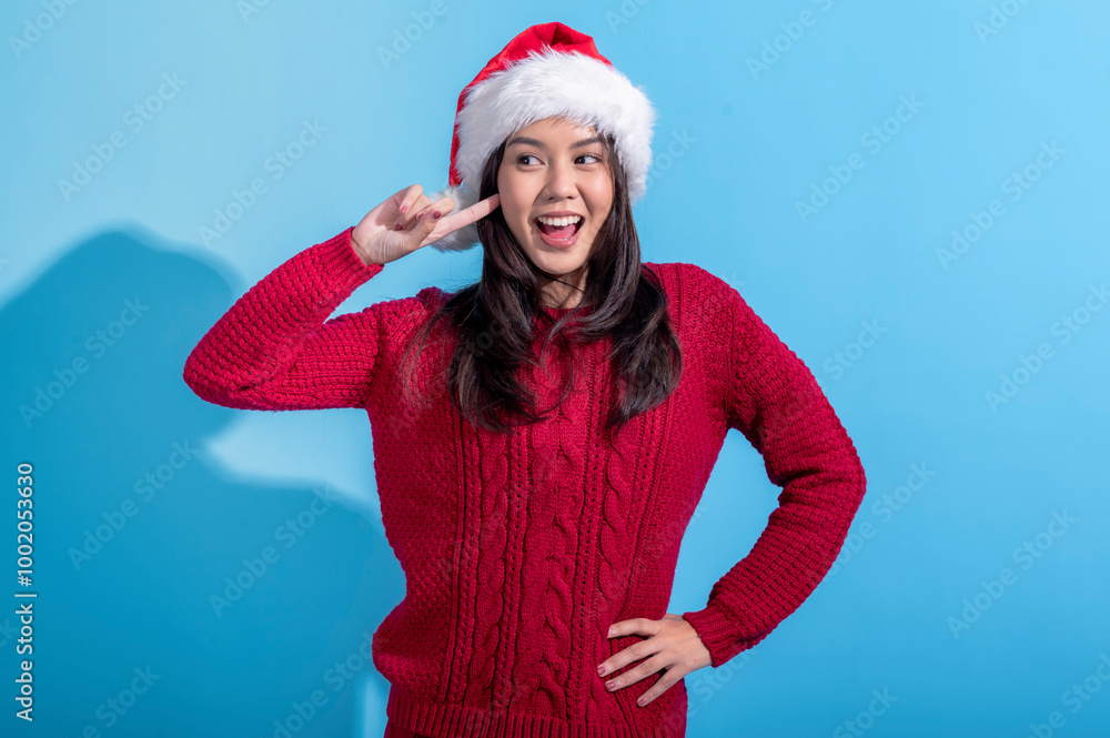 An Asian woman dressed in a red knit sweater and Santa hat playfully posing with a peace sign, smiling brightly. She stands against a light blue background, celebrating the festive Christmas season. An Asian woman dressed in a red knit sweater and Santa hat playfully posing with a peace sign, smiling brightly. She stands against a light blue background, celebrating the festive Christmas season.